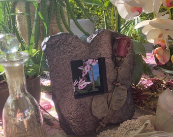May include: A rustic, heart-shaped picture frame with a dried rose and a small picture of pink orchids inside. The frame is sitting on a table with a lace tablecloth and other decorative items.