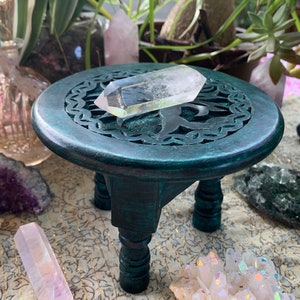 May include: A teal green wooden pedestal with three legs and a carved circular top. A clear quartz crystal point is resting on the top of the pedestal.