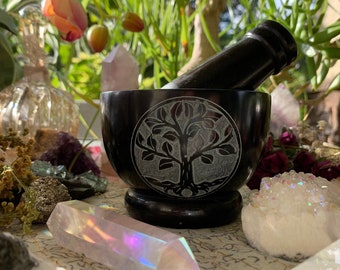 May include: Black stone mortar and pestle with a tree of life design. The mortar is sitting on a table with crystals and other decorative items.