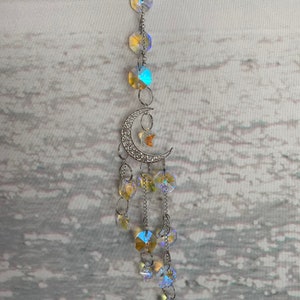May include: A celestial-themed suncatcher featuring a crescent moon and star pendant, adorned with iridescent crystals and linked by a silver chain. The crystals reflect a spectrum of colors, creating a shimmering effect. The suncatcher is designed for hanging.