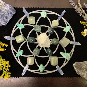 May include: A collection of crystals and stones arranged on a black surface with a white geometric design. The arrangement includes green, blue, and clear stones, along with sprigs of yellow and white flowers and lavender. The crystals vary in size and shape.