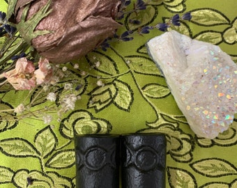 May include: Two black wooden candle holders with a triple moon symbol carved into the front. The holders are sitting on a green floral fabric.