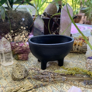 May include: Black cast iron cauldron with three legs, sitting on a table with various crystals, dried flowers, and sage bundles.