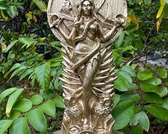 May include: A gold-colored statue of a female figure with a snake wrapped around her. The figure is standing on a base with a circular design. The statue is surrounded by a leafy green background.