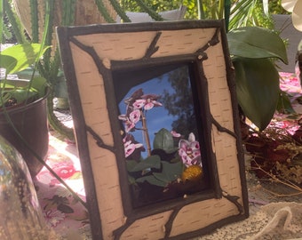 May include: A small, rectangular picture frame made of birch bark with a dark brown border. The frame is sitting on a table with a floral tablecloth and has a photo inside of pink orchids.