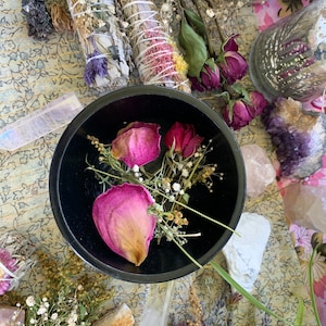 May include: A black bowl filled with dried pink rose petals and white flowers. The bowl is surrounded by various crystals and dried flowers.