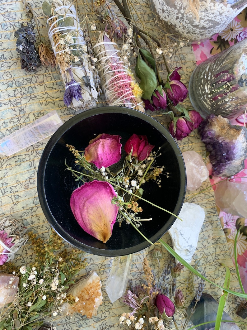 May include: A black bowl filled with dried pink rose petals and white flowers. The bowl is surrounded by various crystals and dried flowers.
