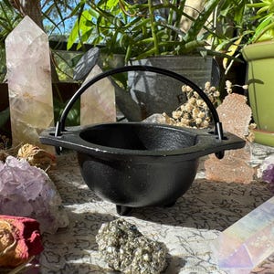 May include: A small, black cast iron cauldron with a curved handle. The cauldron sits on three small feet. The background includes crystals, plants, and other decorative items. The cauldron is a symbol of witchcraft and magic.