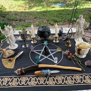 May include: A black altar cloth with gold trim features a pentagram with crystals and a cauldron. A wooden wand, a small broom, and a silver chalice are also on the cloth. There are two white statues of figures and a white marble mortar and pestle on the table.