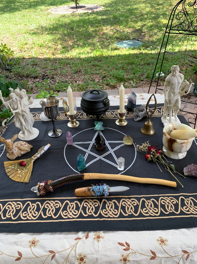 May include: A black altar cloth with gold trim features a pentagram with crystals and a cauldron. A wooden wand, a small broom, and a silver chalice are also on the cloth. There are two white statues of figures and a white marble mortar and pestle on the table.