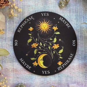 May include: A black round divination board with a sun and moon design. The sun has a smiling face and is surrounded by rays. The moon has a smiling face. The words "Yes", "Maybe", "No", and "Rephrase" are printed around the edge.