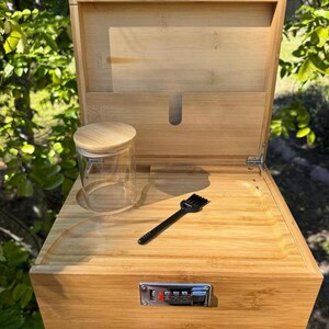 May include: A wooden box with a combination lock, a small glass jar, and a black brush. The box is made of light brown wood and has a hinged lid. The lid is open and the inside of the box is visible. The box is sitting on a green grassy surface.