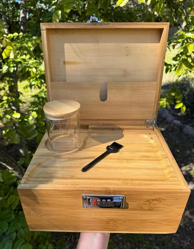 May include: A wooden box with a combination lock, a small glass jar, and a black brush. The box is made of light brown wood and has a hinged lid. The lid is open and the inside of the box is visible. The box is sitting on a green grassy surface.