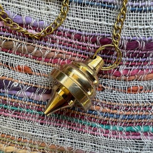 May include: A polished brass pendulum with a tiered, conical shape suspended from a matching gold-toned metal chain. The pendulum features a smooth, reflective surface with a pointed tip and a circular ring at the top for attachment to the chain.