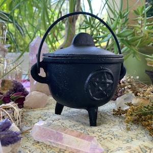 May include: Black cast iron cauldron with a pentagram design and a lid with a handle. The cauldron is sitting on a table with crystals and other items.