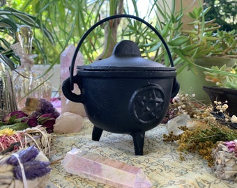 May include: Black cast iron cauldron with a pentagram design and a lid with a handle. The cauldron is sitting on a table with crystals and other items.