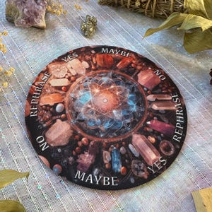 May include: A round divination board with a central mandala design and the words "YES", "NO", "MAYBE", and "REPHRASE" around the edge. The board is decorated with various crystals in shades of red, brown, and blue.