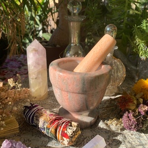 May include: A pink and gray marble mortar and pestle with a wooden pestle. The mortar and pestle are surrounded by dried flowers, crystals, and a smudge stick.