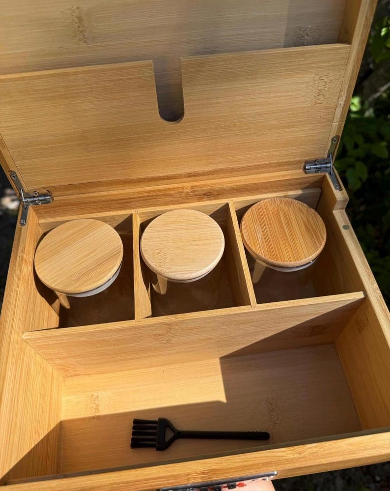 May include: A wooden box with a hinged lid, containing three small glass jars with wooden lids. The box has a small compartment for a brush, which is included.