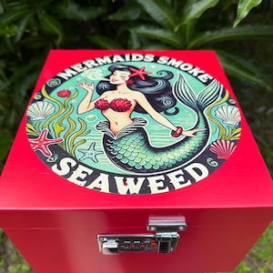 May include: A bright red wooden box with a combination lock on the front. The top features a circular graphic of a mermaid with long black hair, a green tail, and red seashell bra, surrounded by seaweed and shells, with the text MERMAIDS SMOKE SEAWEED.