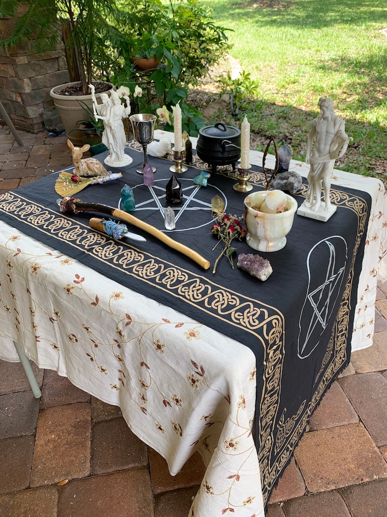 May include: A black altar cloth with gold trim and a pentagram design. The cloth is laid out on a white tablecloth with floral embroidery. The altar is decorated with candles, crystals, statues, and other ritual objects.