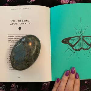 May include: An open book with a teal cover and a black and white illustration of a butterfly. The book is titled "Spell to Bring About Change". A green stone is resting on the open pages of the book.