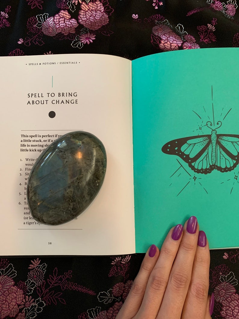 May include: An open book with a teal cover and a black and white illustration of a butterfly. The book is titled "Spell to Bring About Change". A green stone is resting on the open pages of the book.