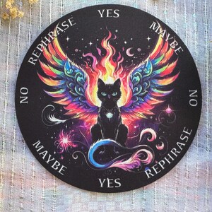 May include: A black round divination mat featuring a black cat with colorful phoenix wings and flames. The mat has the words "Yes", "No", "Maybe", and "Rephrase" around the edge. The cat has a jewel on its chest.