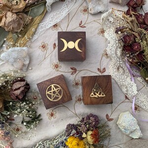May include: Three small, dark wooden boxes with gold-colored symbols: a triple moon, a pentagram, and a Celtic knot. The boxes are arranged on a floral patterned cloth, surrounded by dried flowers and crystals.