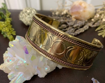 Könnte beinhalten: Ein breites, goldfarbenes Armband mit einem sich wiederholenden Halbmond-Design. Das Armband hat eine geflochtene, kupferfarbene Bordüre. Der Schmuck wird auf einem weißen Kristallcluster präsentiert.