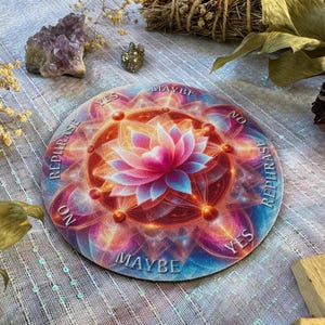 May include: A round divination board with a lotus flower design in shades of pink, orange, and blue. The words "YES", "NO", "MAYBE", and "REPHRASE" are printed around the edge. The board is surrounded by crystals and dried plants.