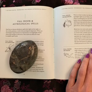 May include: An open book with a grey stone on the left page and a hand holding the book on the right page. The book is titled "Full Moon & Astrological Spells". The text on the left page reads "Capricorn brings goals no" and "highlights which make things honest about". The text on the right page reads "Full Moon in Aquarius" and "This is a time to allow the full moon spotlight right on you! Make time for what is working for you and what habits that are holding you back are shifting energy patterns that are holding you back; make space for new rituals, gain focus, clarity and happiness."
