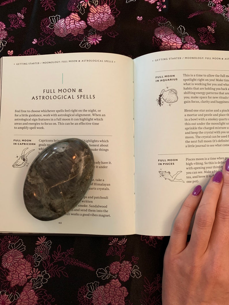 May include: An open book with a grey stone on the left page and a hand holding the book on the right page. The book is titled "Full Moon & Astrological Spells". The text on the left page reads "Capricorn brings goals no" and "highlights which make things honest about". The text on the right page reads "Full Moon in Aquarius" and "This is a time to allow the full moon spotlight right on you! Make time for what is working for you and what habits that are holding you back are shifting energy patterns that are holding you back; make space for new rituals, gain focus, clarity and happiness."