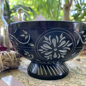May include: A black carved stone bowl with a white floral design. The bowl has a wide, round base and a deep, rounded interior. The bowl is perfect for holding incense, crystals, or other small items.