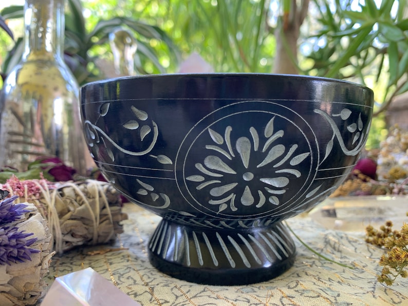 May include: A black carved stone bowl with a white floral design. The bowl has a wide, round base and a deep, rounded interior. The bowl is perfect for holding incense, crystals, or other small items.