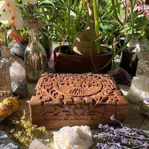 May include: A wooden box with intricate carvings of a tree of life design. The box is brown and has a hinged lid. The box is surrounded by crystals, dried flowers, and other natural elements.