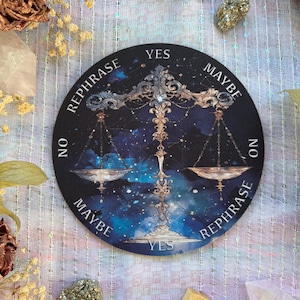 May include: A round, black divination board featuring a celestial design with a scale illustration. The board has the words "YES", "MAYBE", "NO", and "REPHRASE" around the edges. The background is a starry night sky with blue and purple hues.