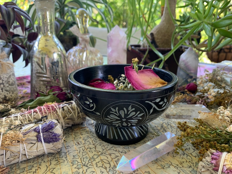 May include: A black carved bowl with dried rose petals and white flowers inside. The bowl is on a table with other items, including a bundle of sage, a clear glass bottle, and a crystal.