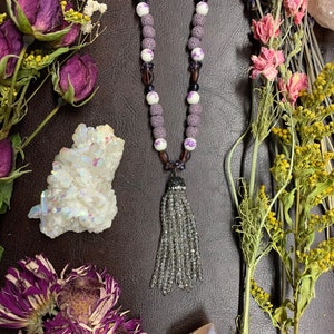 May include: A purple and white beaded necklace with a black tassel pendant. The necklace is made of lava rock beads and has a floral pattern. The necklace is surrounded by dried flowers and crystals.