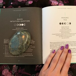 May include: An open book with a green cover and white pages. The book is titled "Spells & Potions: Third Eye" and features two different spells: "Quick Intuition Question" and "Psychic Tea". The book is open to a page with instructions for each spell, including the ingredients needed. A hand with purple nail polish is holding the book open.