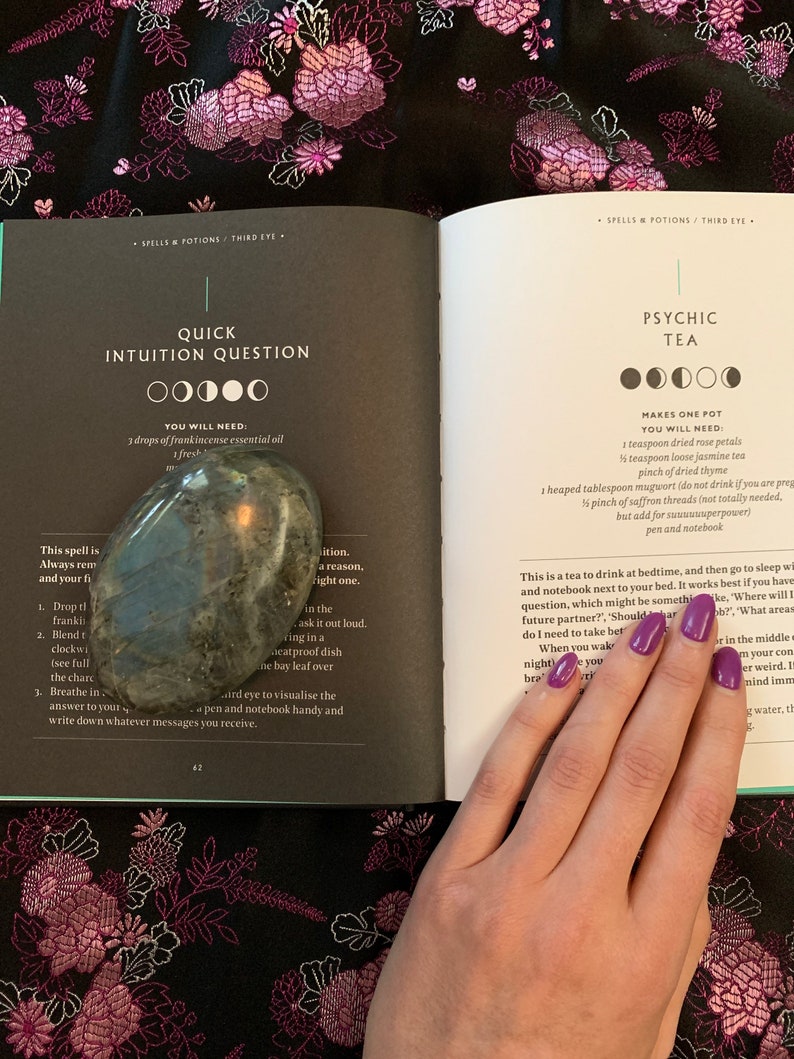 May include: An open book with a green cover and white pages. The book is titled "Spells & Potions: Third Eye" and features two different spells: "Quick Intuition Question" and "Psychic Tea". The book is open to a page with instructions for each spell, including the ingredients needed. A hand with purple nail polish is holding the book open.