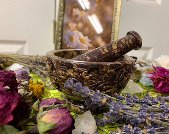 May include: A brown and black stone mortar and pestle with intricate carvings. The mortar and pestle are surrounded by dried flowers and crystals.