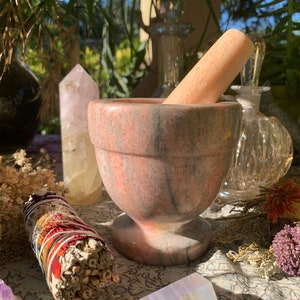 May include: A pink and gray marble mortar and pestle set. The mortar has a wide, round base and a deep bowl. The pestle is a long, cylindrical shape. The set is sitting on a table with other items, including dried flowers and crystals.