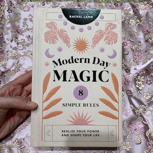 May include: A book titled "Modern Day Magic" by Rachel Lang. The cover features a cream background with illustrations of plants, suns, moons, and stars in shades of pink, orange, and purple. The text "8 Simple Rules" and "Realize Your Power and Shape Your Life" are also visible.