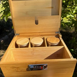 May include: A wooden box with a combination lock and three glass jars inside. The box is made of light brown wood and has a natural finish. The jars are clear glass and have lids made of the same wood as the box.