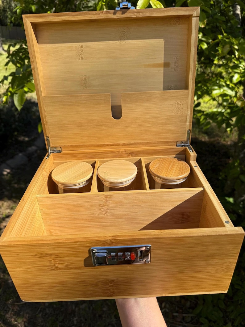 May include: A wooden box with a combination lock and three glass jars inside. The box is made of light brown wood and has a natural finish. The jars are clear glass and have lids made of the same wood as the box.