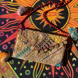 May include: A gold and cream patchwork clutch bag with a beaded tassel and coin accents. The bag is on a colorful tapestry with a sun design.