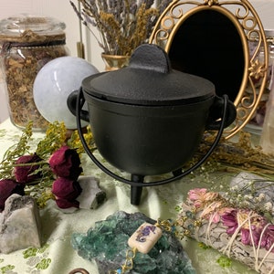 May include: A black cast iron cauldron with a lid, set on a table with dried flowers, crystals, and a decorative key. A gold-framed mirror and a jar filled with herbs are in the background. The cauldron has a handle on each side.