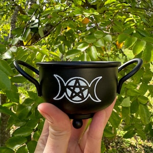 May include: A small, black cauldron with two handles and a decorative base. The cauldron features a white triple moon symbol with a pentagram in the center. The background is a blurred view of green foliage.