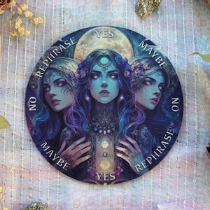 May include: Round divination board featuring three ethereal women with blue skin and flowing hair, surrounded by a crescent moon. The board has the words "Yes", "Maybe", "No", and "Rephrase" around the edges. The artwork is in shades of blue, purple, and white.